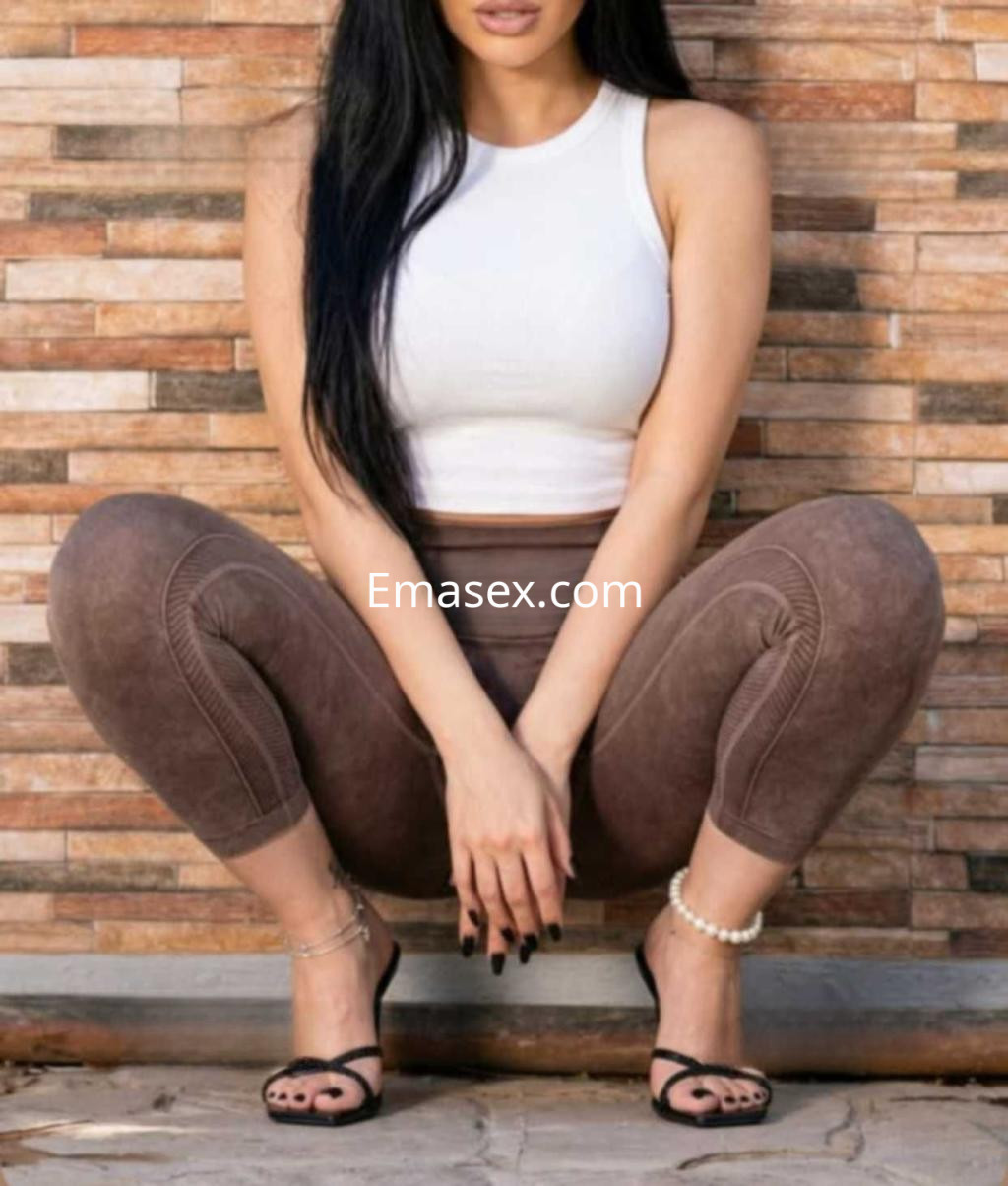 Puta en Castellón luciendo una blusa blanca sin mangas y leggings marrones, posando en cuclillas sobre un fondo de piedra.