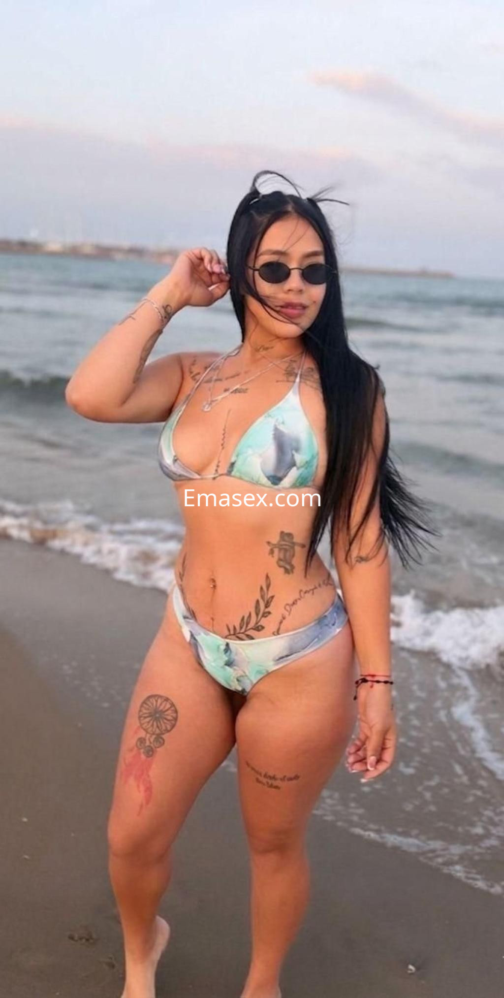 Escort en la playa de Castellón, luciendo un bikini de estampado floral y gafas de sol, con tatuajes visibles.