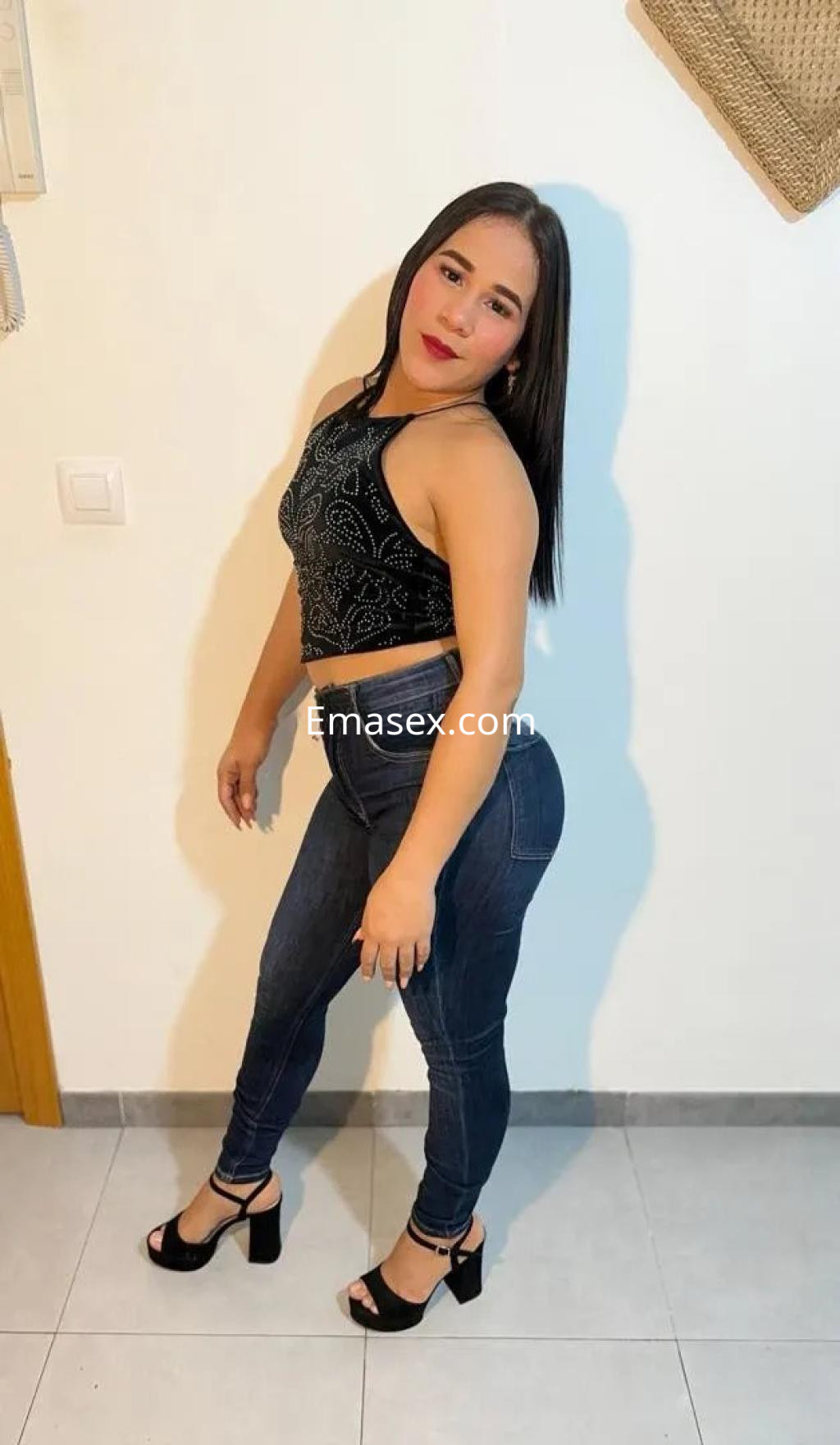 Escort en Córdoba con blusa negra sin mangas, jeans ajustados y tacones, posando sonriente en un ambiente interior.