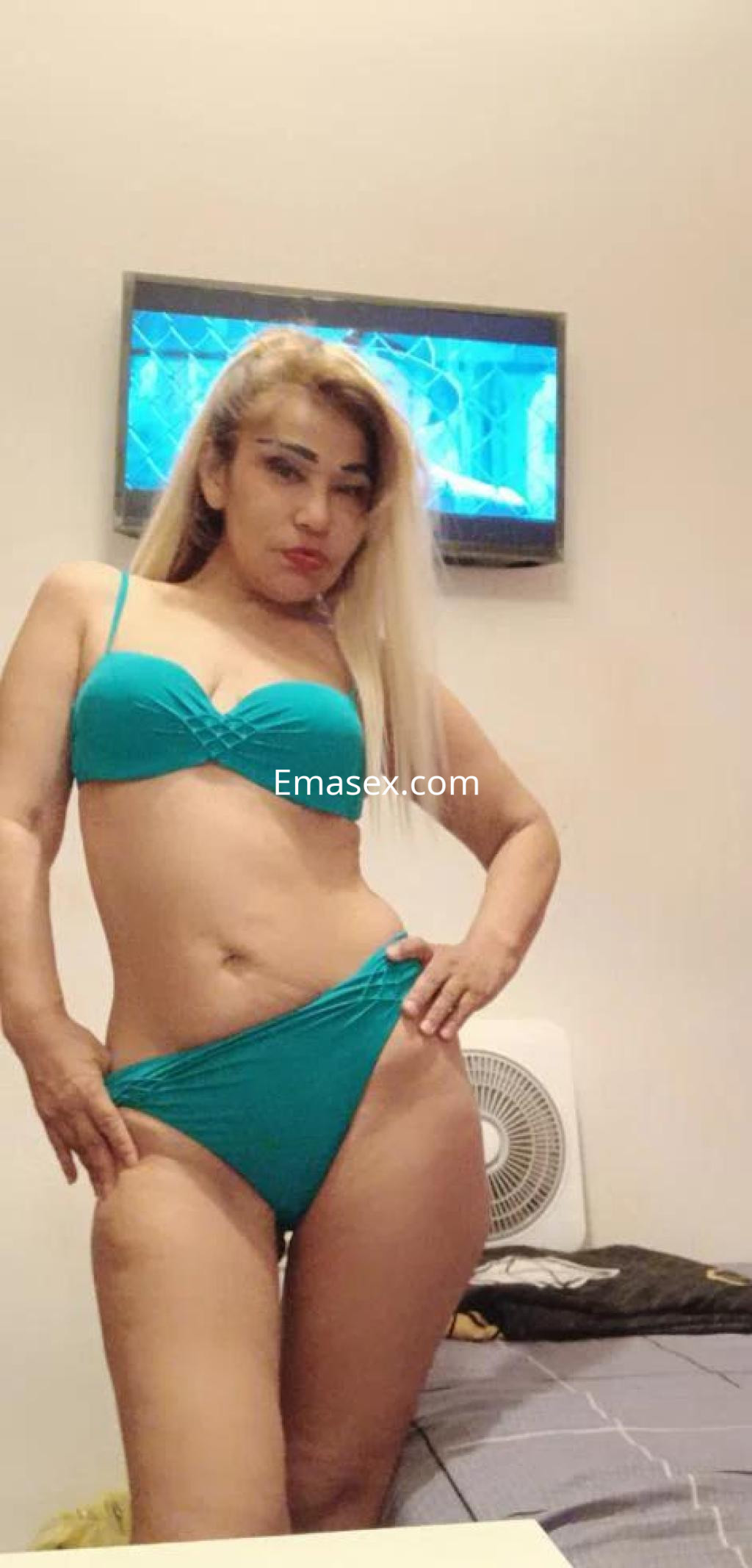 Escort en Madrid, posando con un bikini verde, cabello rubio y lacio, en un ambiente interior.