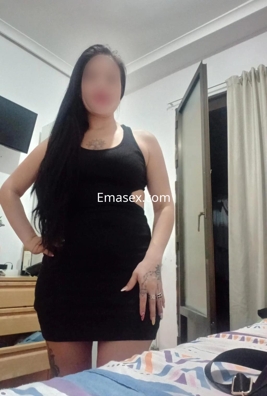 Escort en Murcia con vestido negro ajustado y cabello largo, posando en una habitación con luz natural.