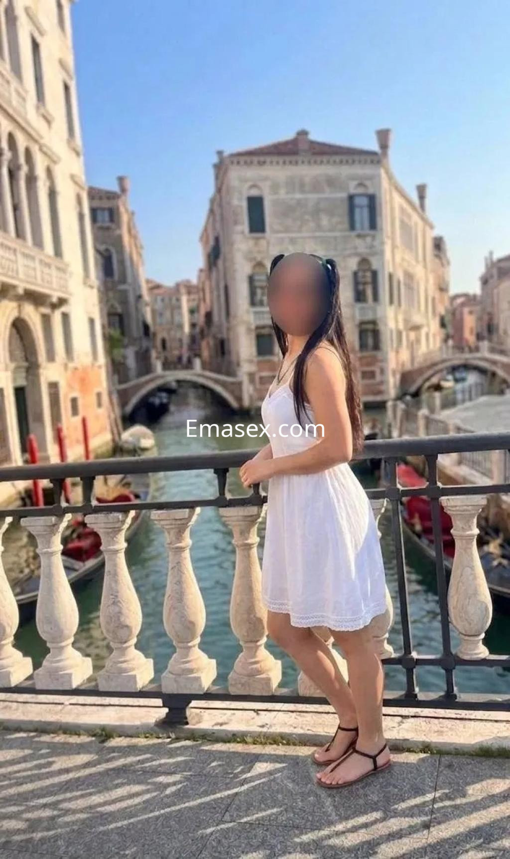 Escort en Valencia con vestido blanco, posando junto a un canal y edificios históricos en un día soleado.