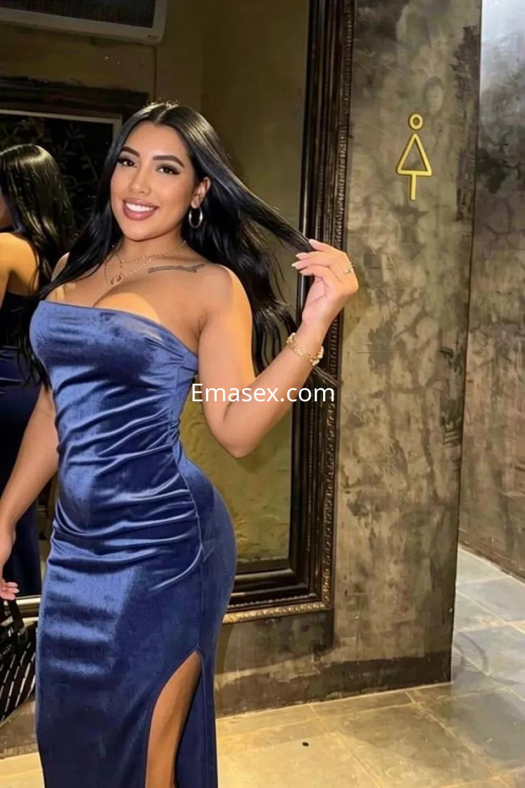 Escort en Valencia luciendo un vestido azul ajustado con abertura lateral, sonriendo y posando frente a un espejo.