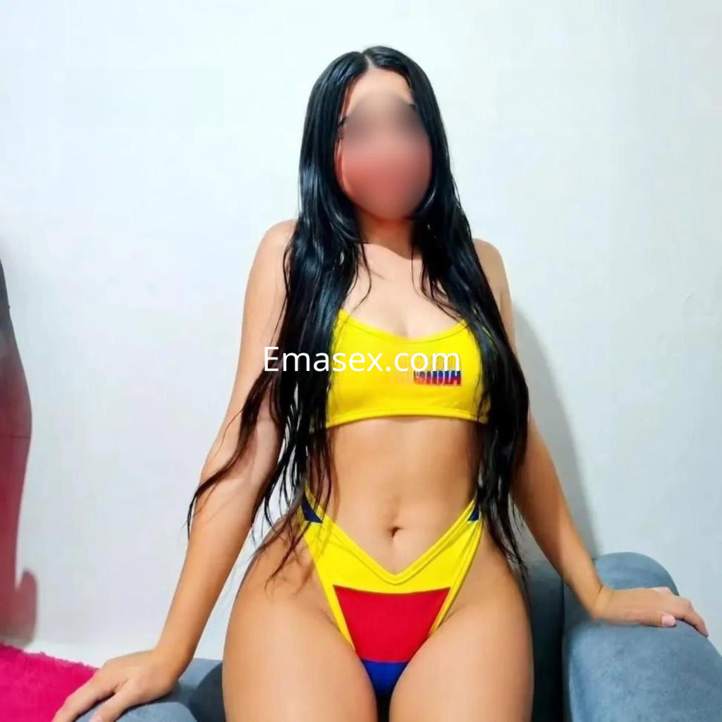 Escort en Valladolid luce un bikini amarillo con detalles en rojo y azul, mostrando una pose relajada en un sofá.