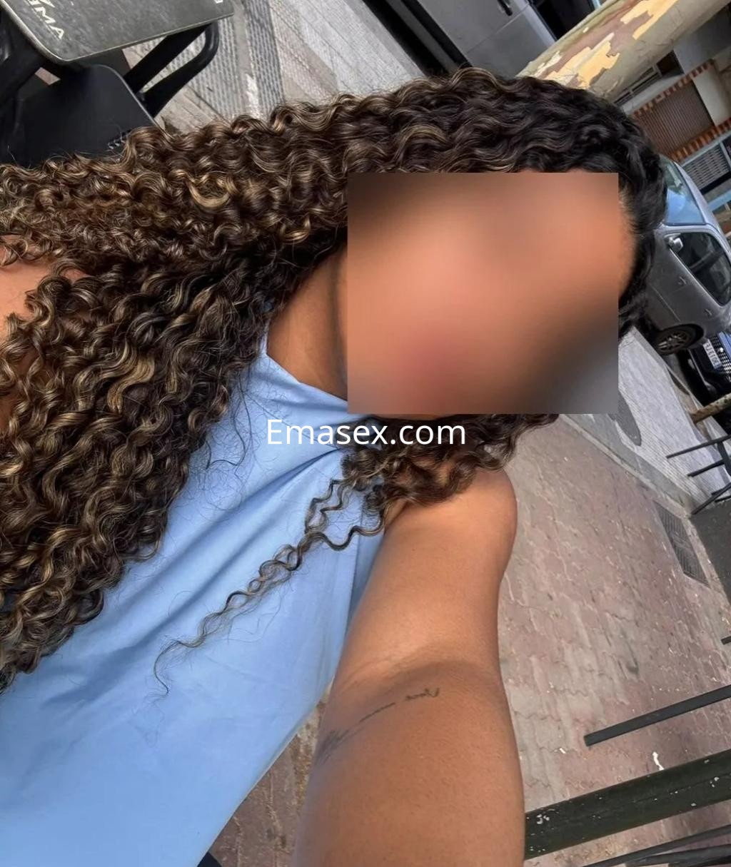 La puta aparece con cabello rizado y suelto, vistiendo una blusa azul, en un entorno urbano de Zaragoza.