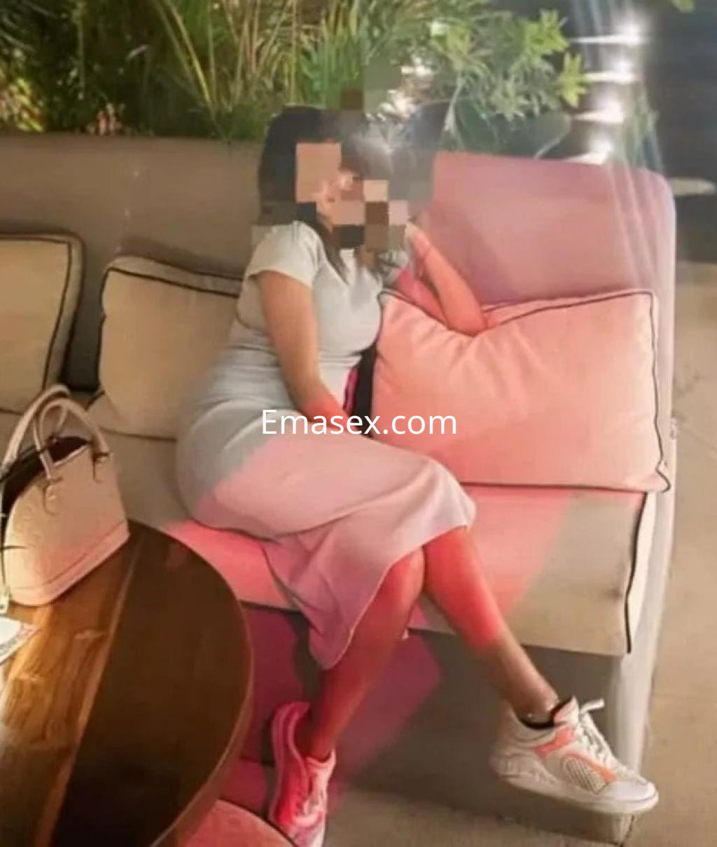 Escort en Zaragoza lleva un vestido claro, sentada en un sofá con una mano en la cabeza y zapatillas deportivas.