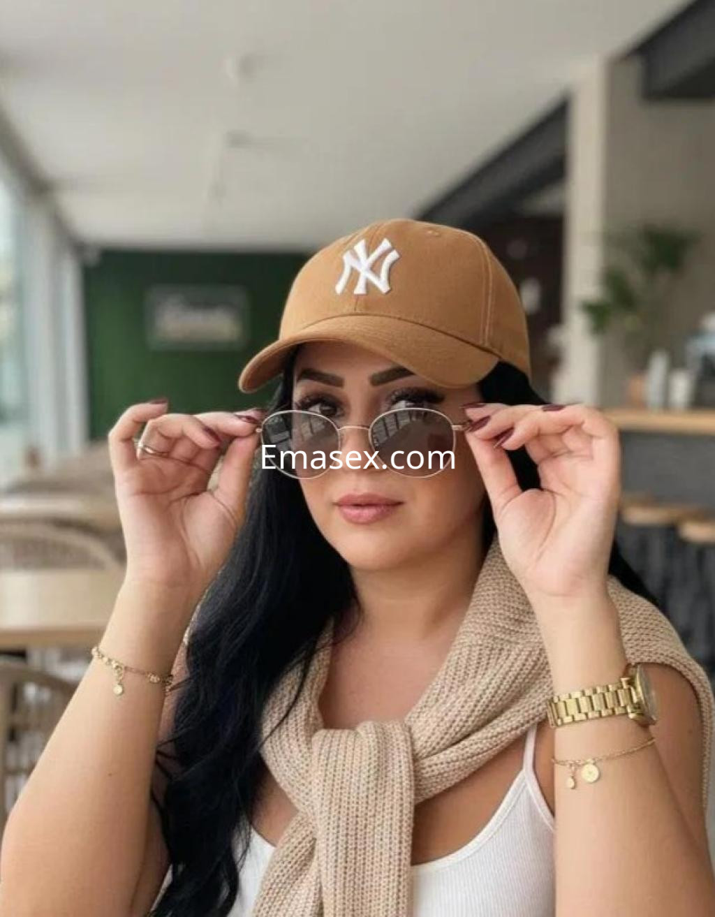 Puta en Alicante, con gorra marrón y gafas de sol, viste una blusa blanca y un suéter atado al cuello.