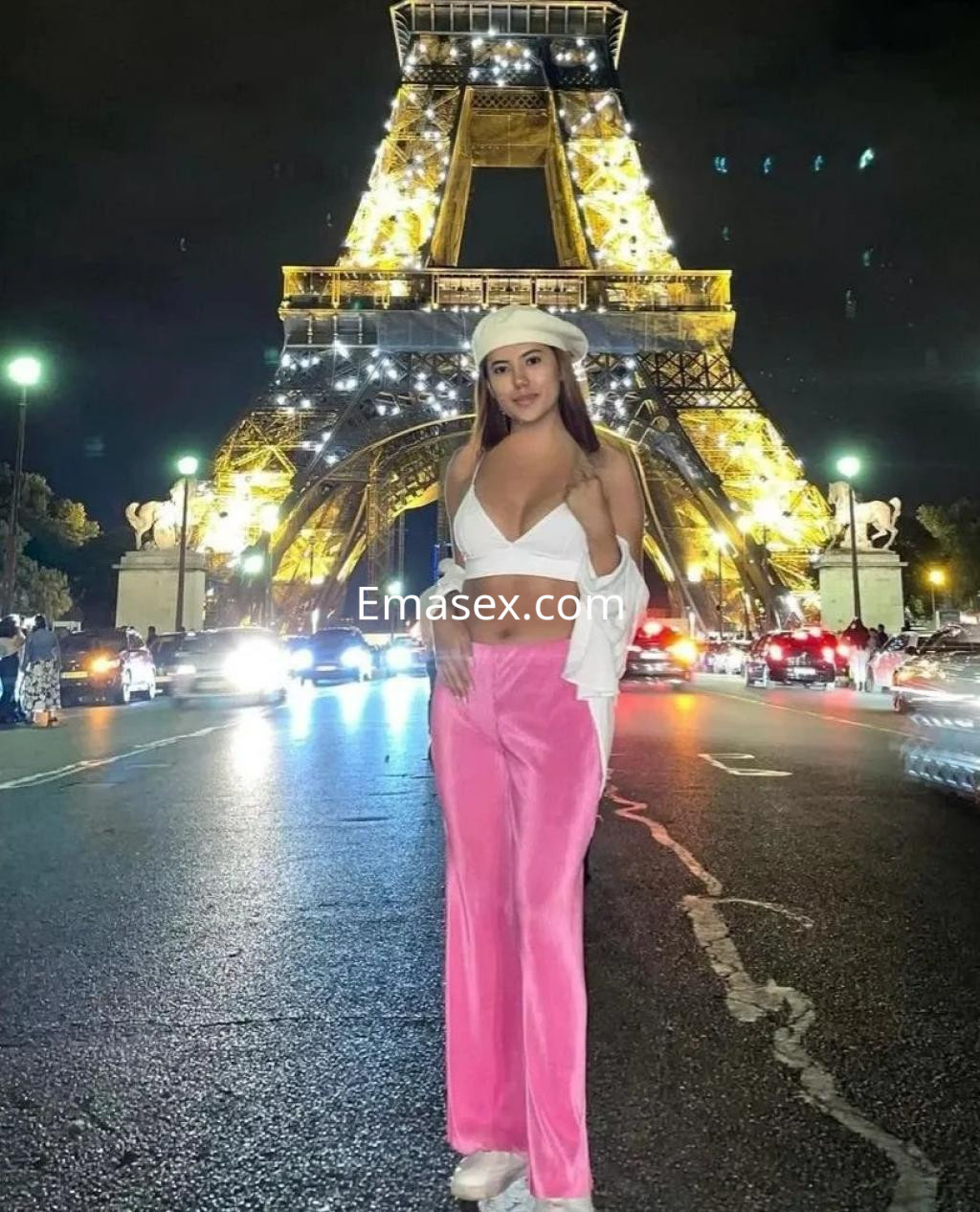 Puta en Alicante con cabello lacio, viste un top blanco y pantalones rosas, posando frente a la Torre Eiffel iluminada.