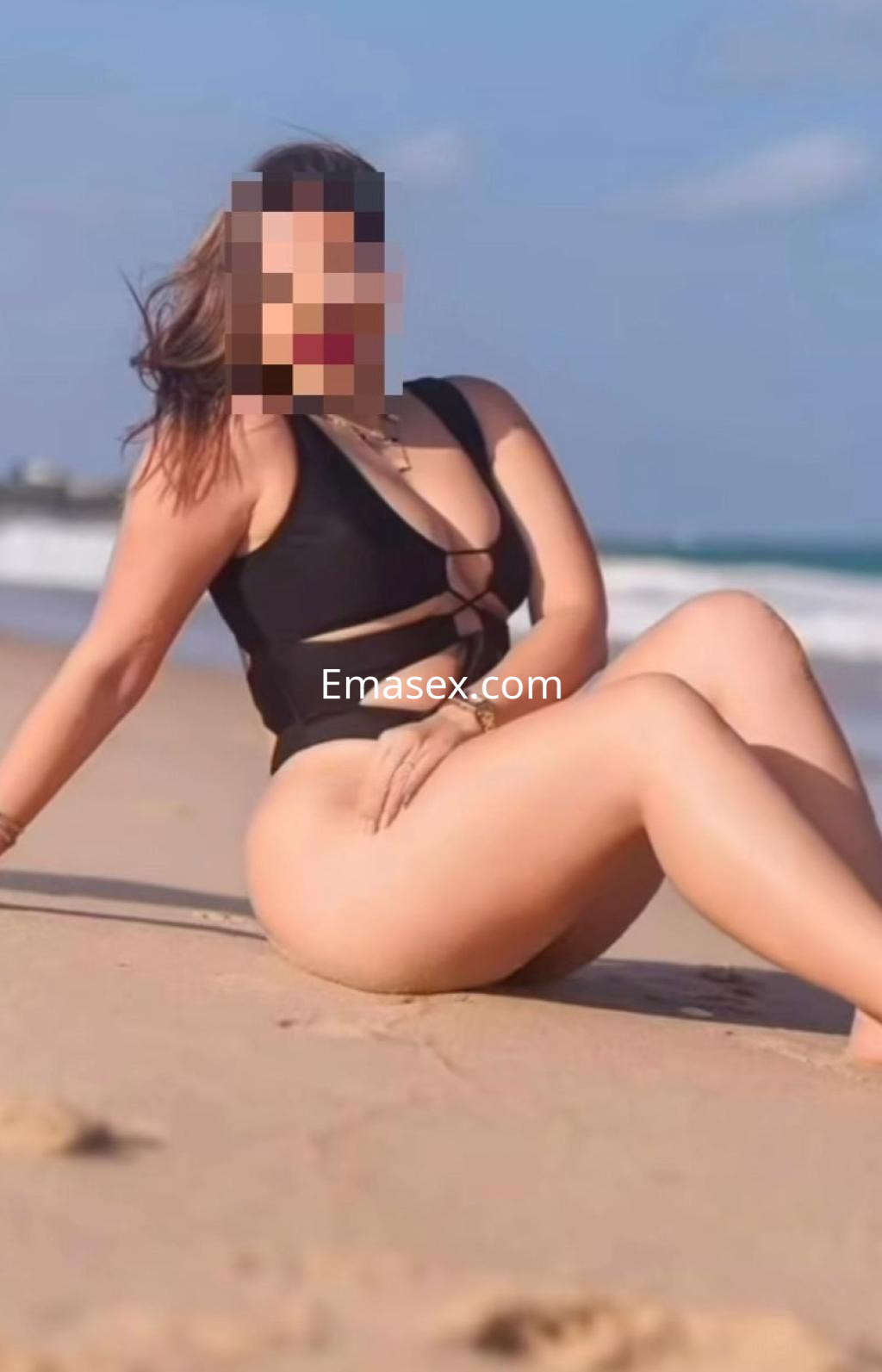 Puta en la playa de Cádiz, luciendo un bañador negro, sentada sobre la arena con una pose relajada.