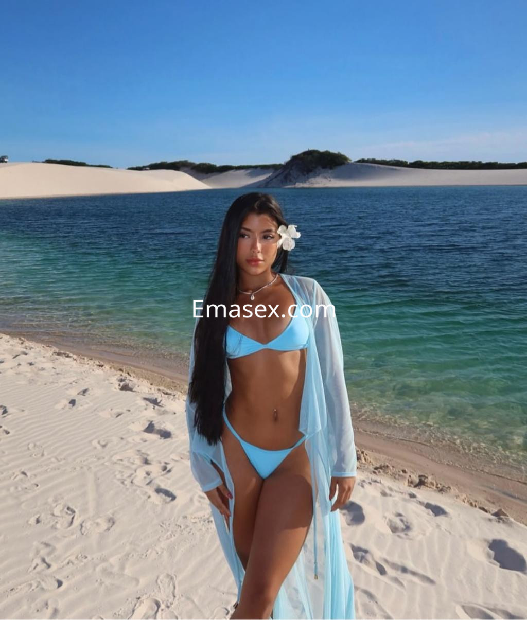 Escort en Málaga con bikini azul y kimono transparente, posando en la playa junto al agua turquesa.