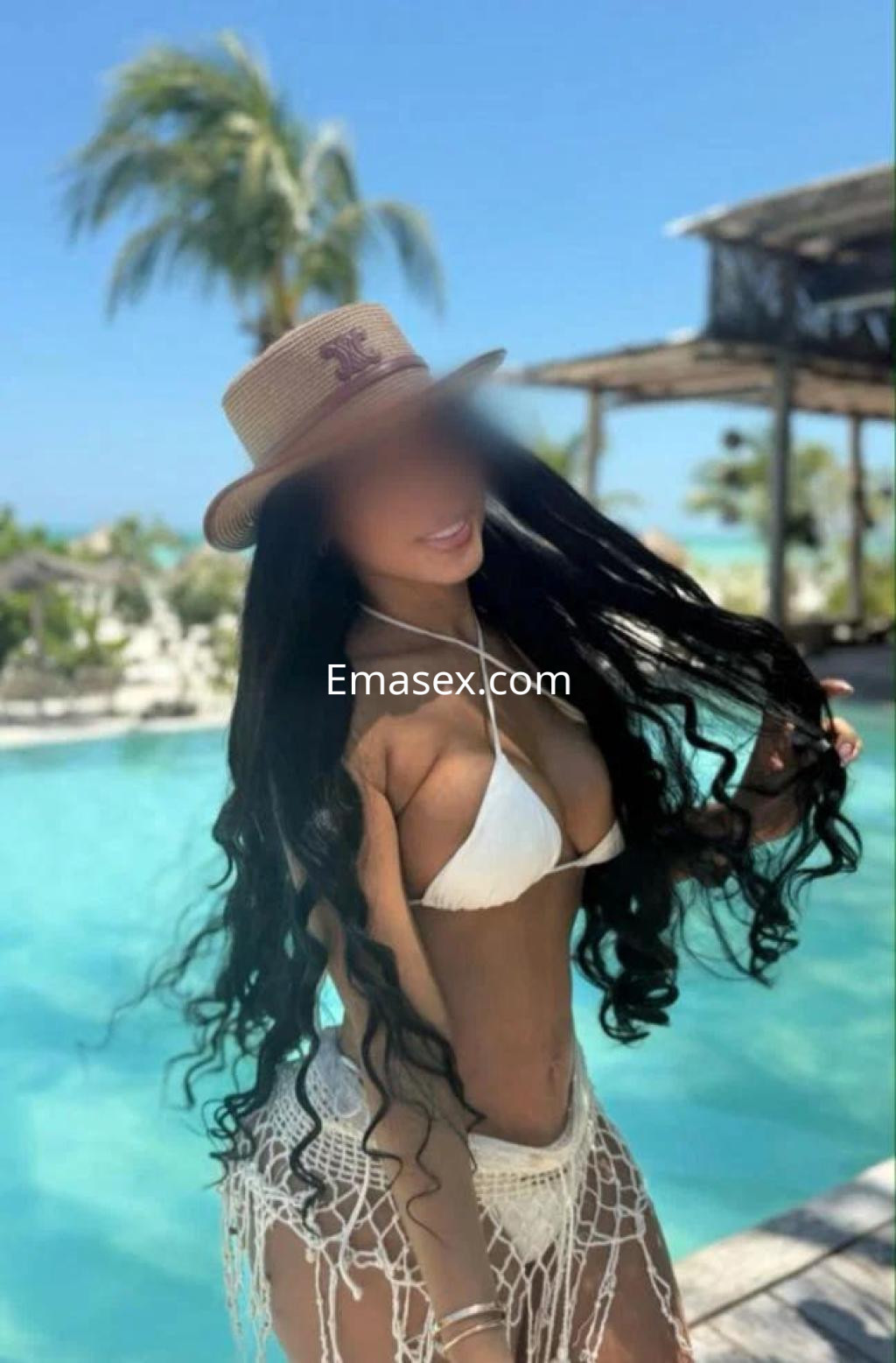 Escort en Albacete con cabello largo y rizado, usando un bikini blanco y sombrero, posando junto a una piscina.