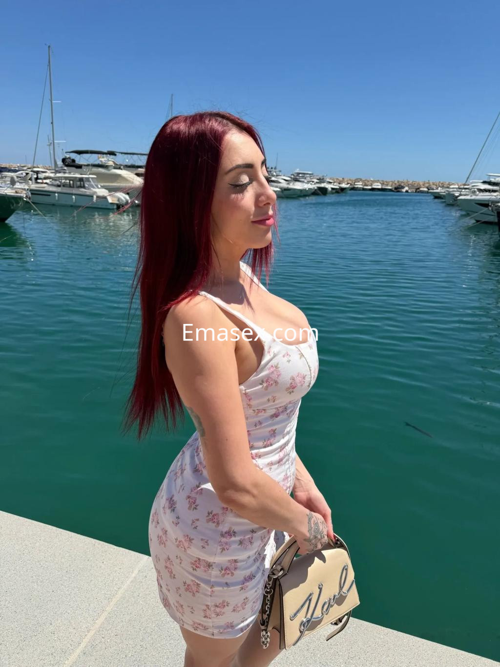 Escort en Palma de Mallorca, luciendo un vestido blanco con estampado floral, junto a un puerto deportivo.
