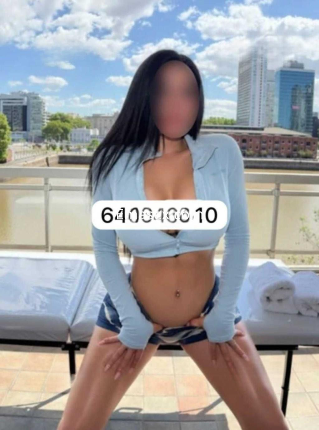 Puta en Barcelona luciendo un top celeste y shorts cortos, posando en una terraza con vista a la ciudad.