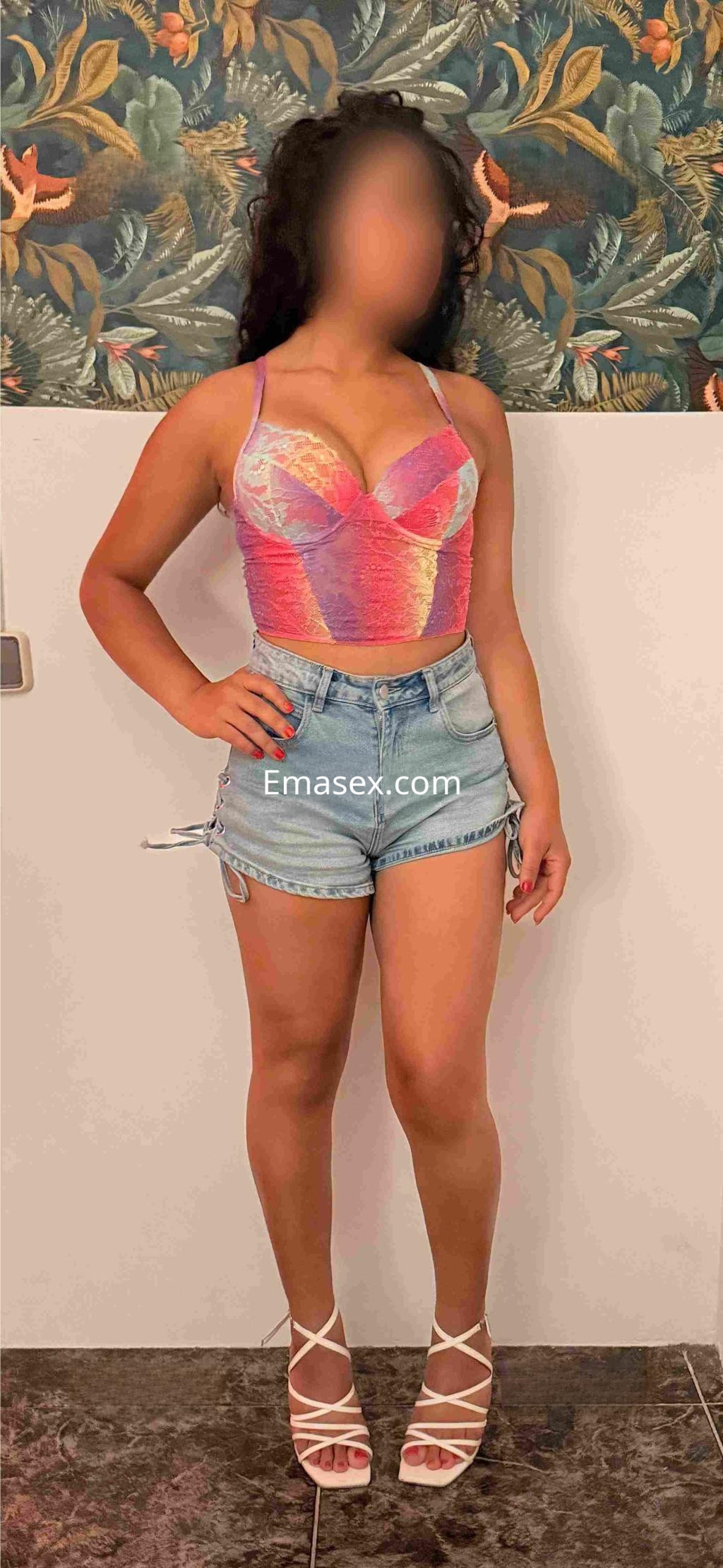 Escort en Barcelona con blusa colorida, shorts de mezclilla y sandalias blancas, posando en un entorno decorativo.