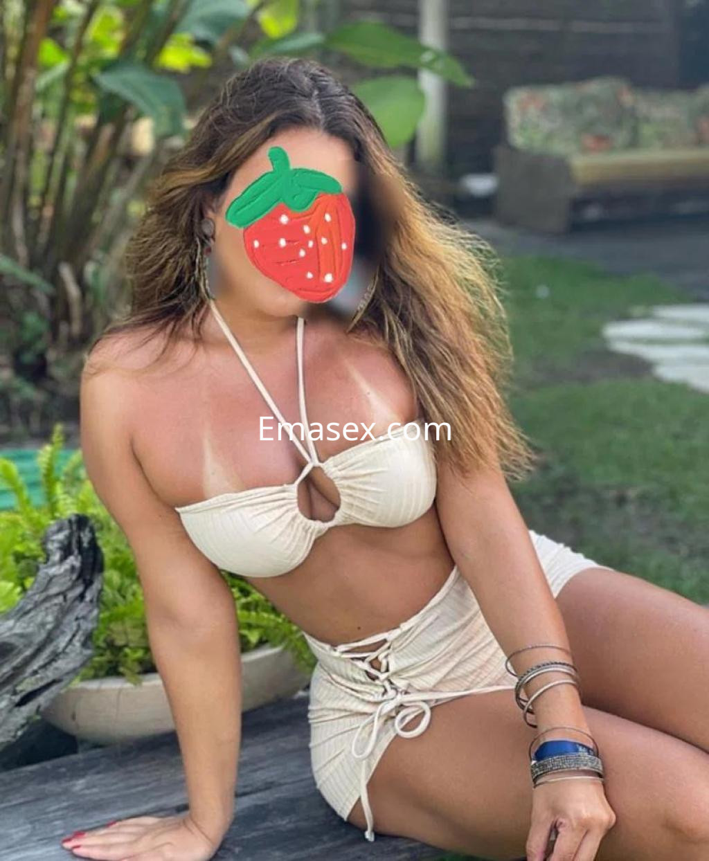 Escort en Cádiz, con cabello ondulado y bronceado, posa en bikini claro en un entorno tropical.