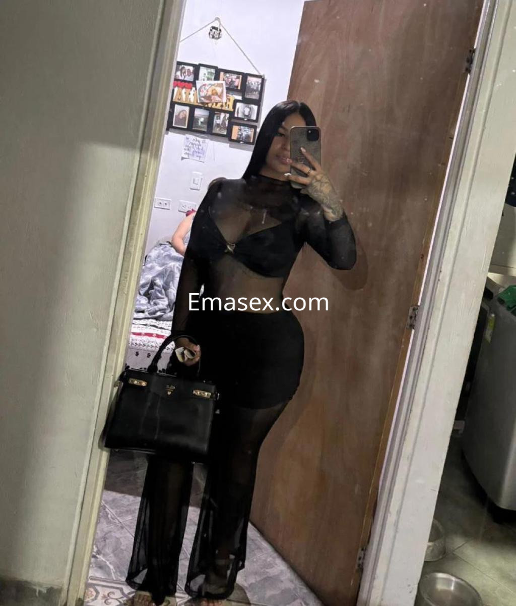 Escort en Madrid de pie frente a un espejo, vistiendo un conjunto negro translúcido y sosteniendo un bolso.