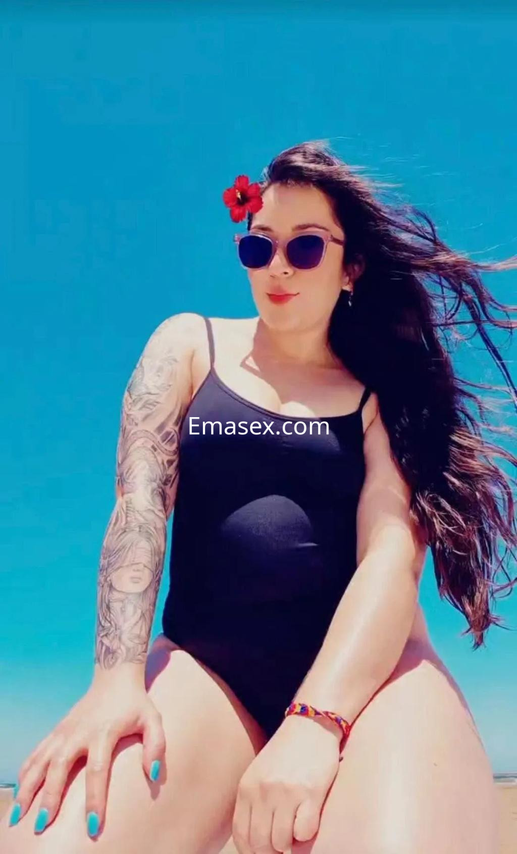 La puta en Gijón posa con un bañador negro, gafas de sol y un tatuaje en el brazo, frente a un cielo azul.