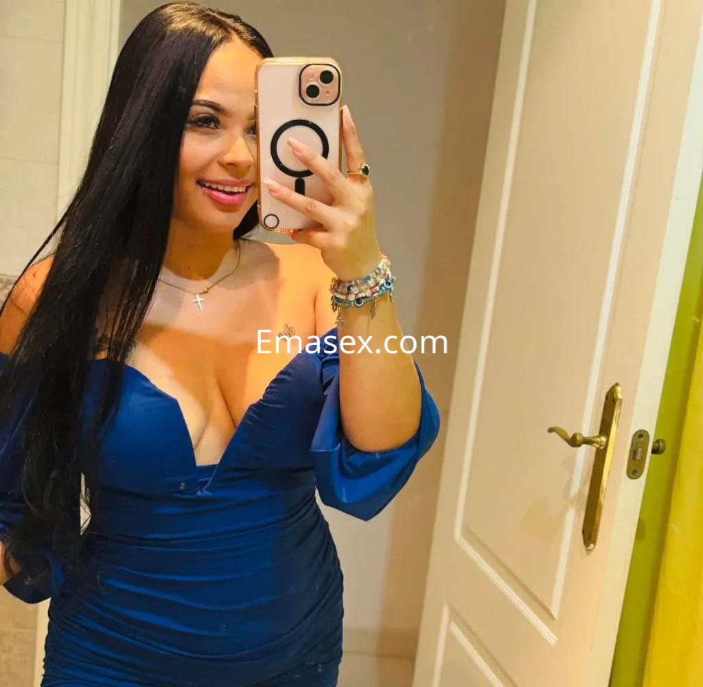 Escort en Valencia con cabello largo y lacio, usando un vestido azul de escote, sonríe mientras toma una selfie.