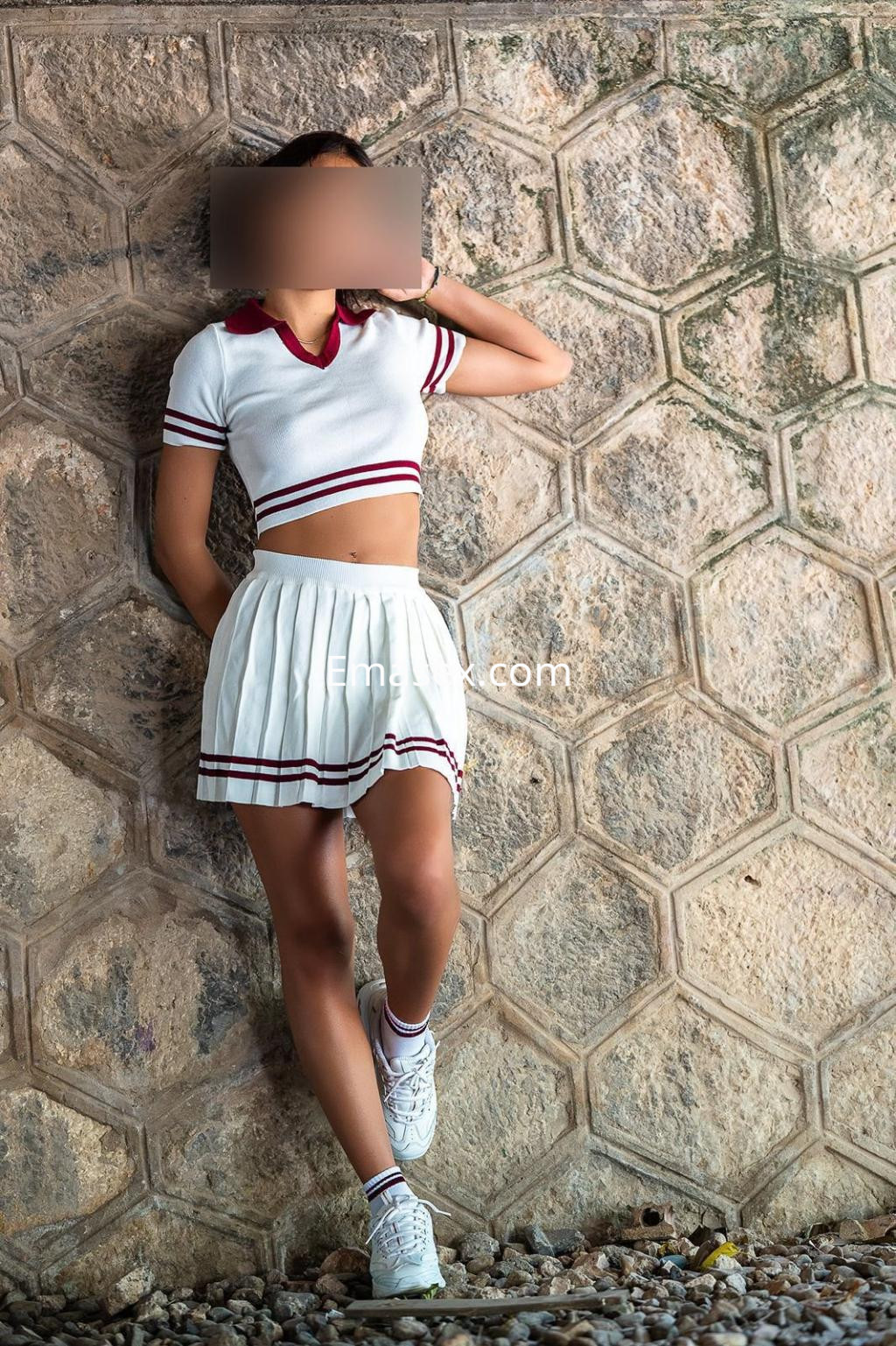 La escort en Valencia viste un conjunto deportivo de falda corta blanca y camiseta con detalles rojos, apoyada en una pared.