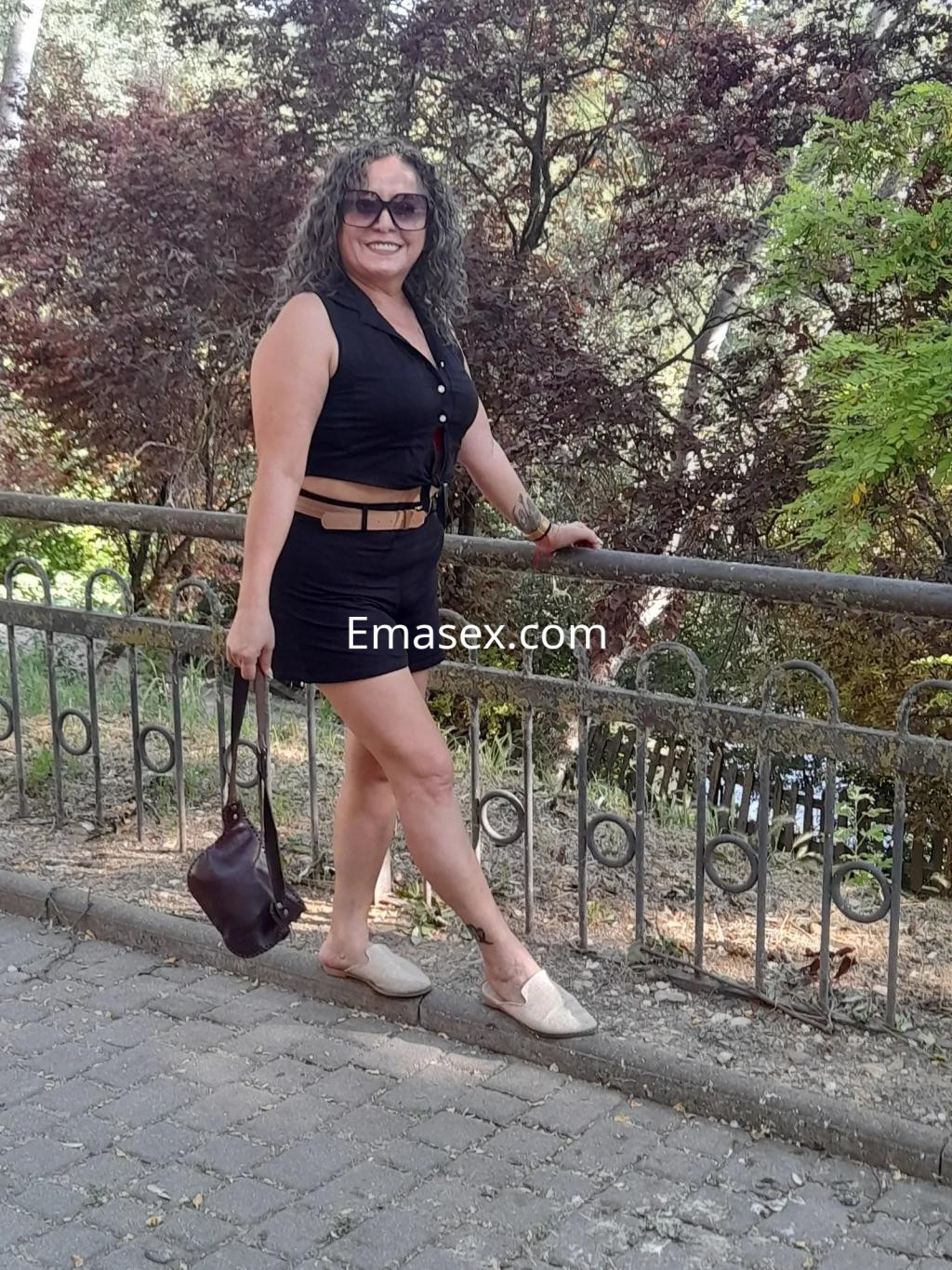 Puta de cabello rizado y oscuro, viste un vestido negro ajustado y sandalias, posando en un parque de Valladolid.
