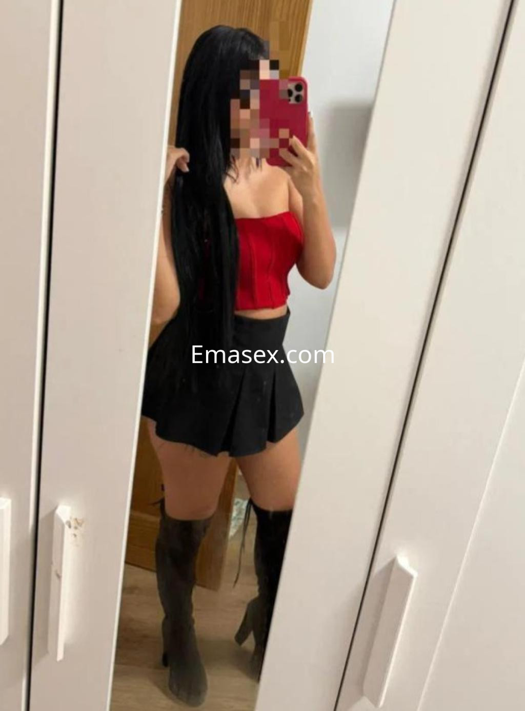 Escort en Sevilla, posando frente a un espejo con top rojo, falda corta negra y botas altas, mostrando su largo cabello.