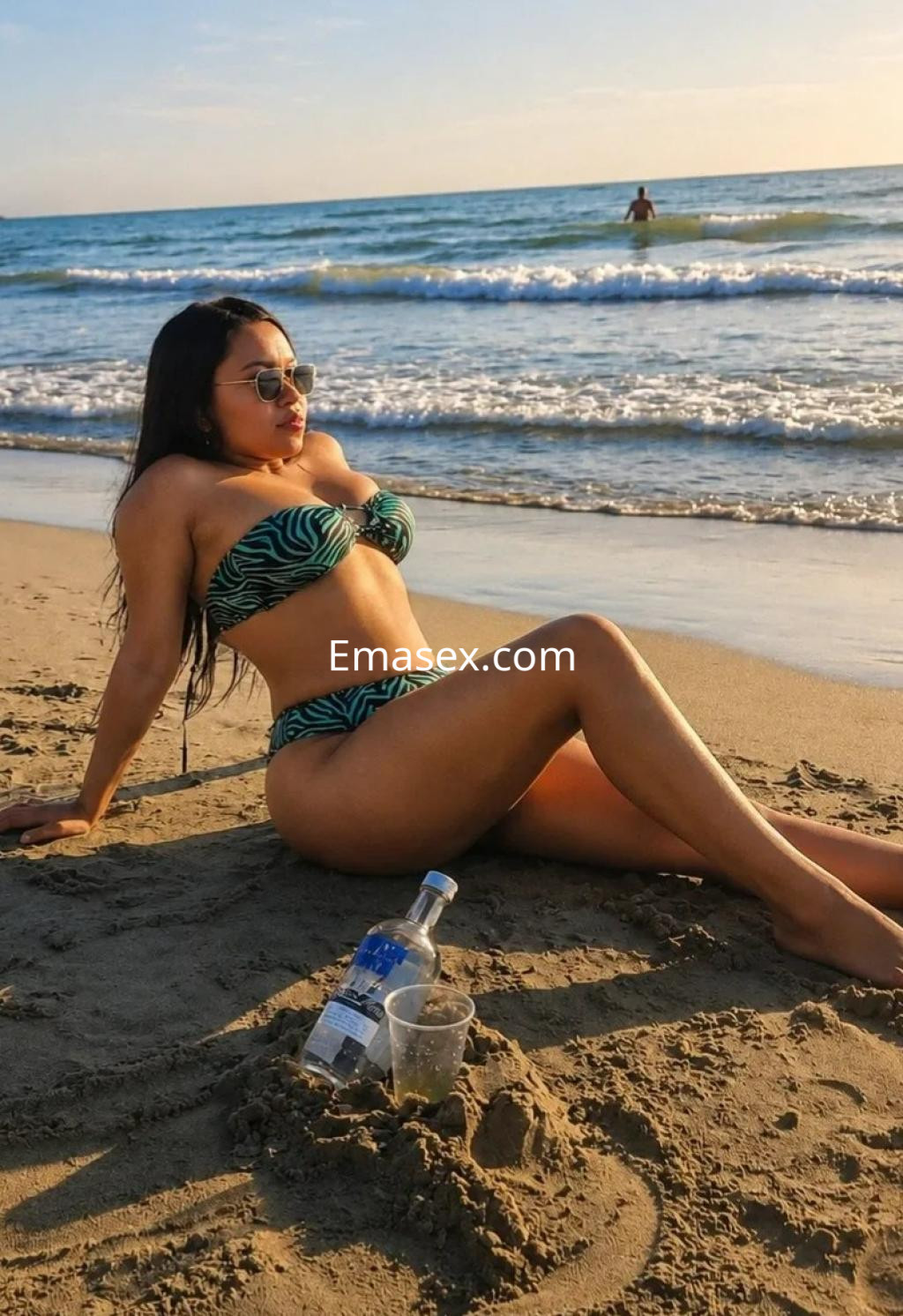 Escort en la playa de Alicante, luciendo un bikini de rayas y gafas de sol, con una botella cerca.