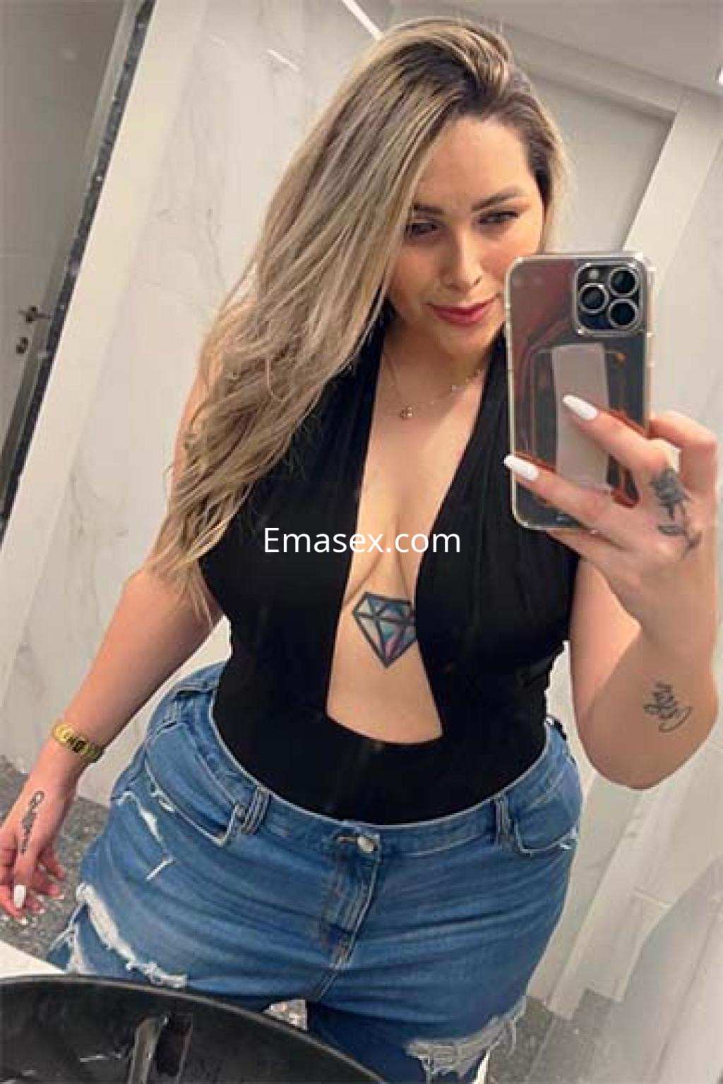 Puta en Logroño posando en un espejo con un body negro y jeans cortos, mostrando un tatuaje en el pecho.