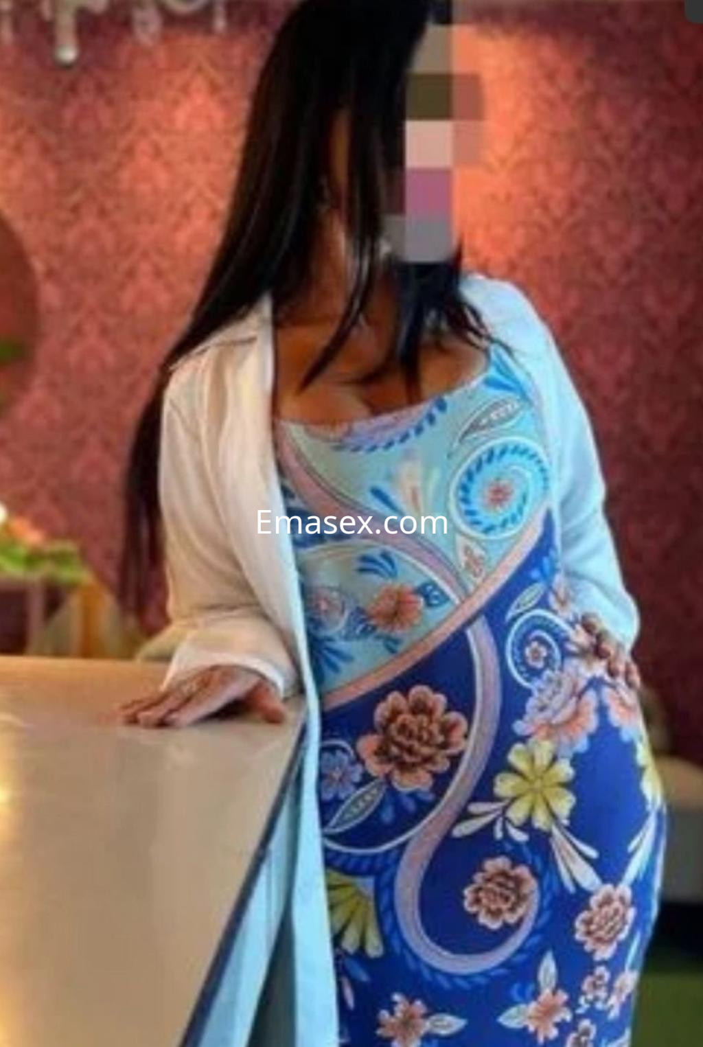 Puta en Cádiz, posando junto a una barra con un vestido colorido de flores y un abrigo blanco.