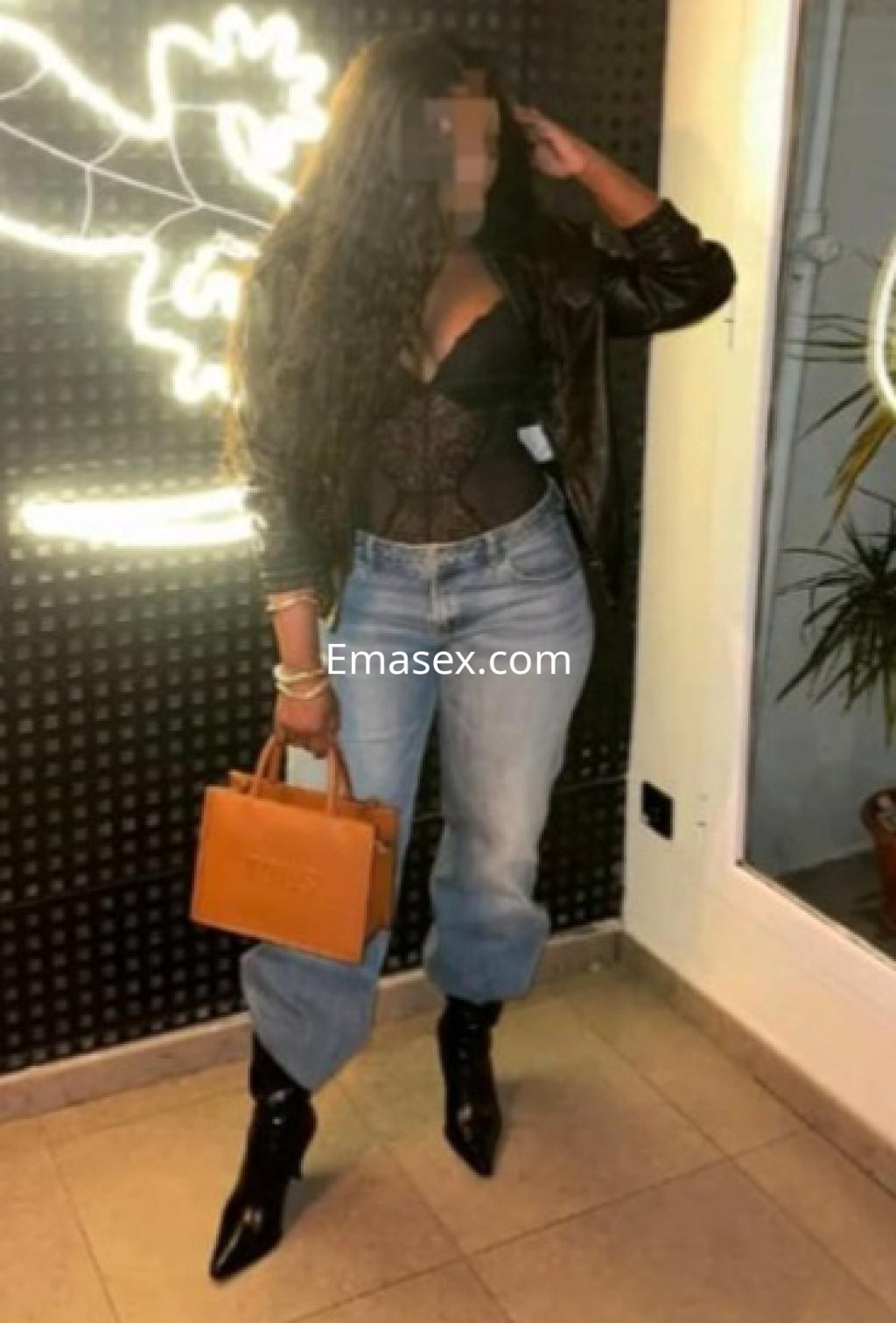 Escort en Cádiz viste un body negro con chaqueta, jeans claros y botas altas, sosteniendo un bolso marrón.