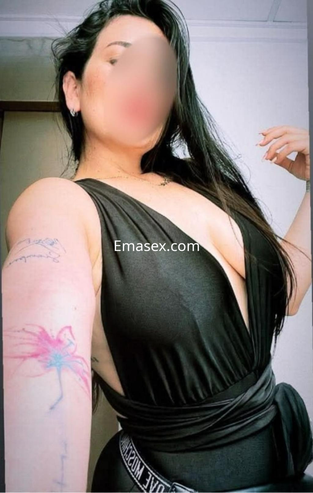 Puta en Valladolid con un vestido negro de escote profundo, mostrando su tatuaje en el brazo y cabello largo.