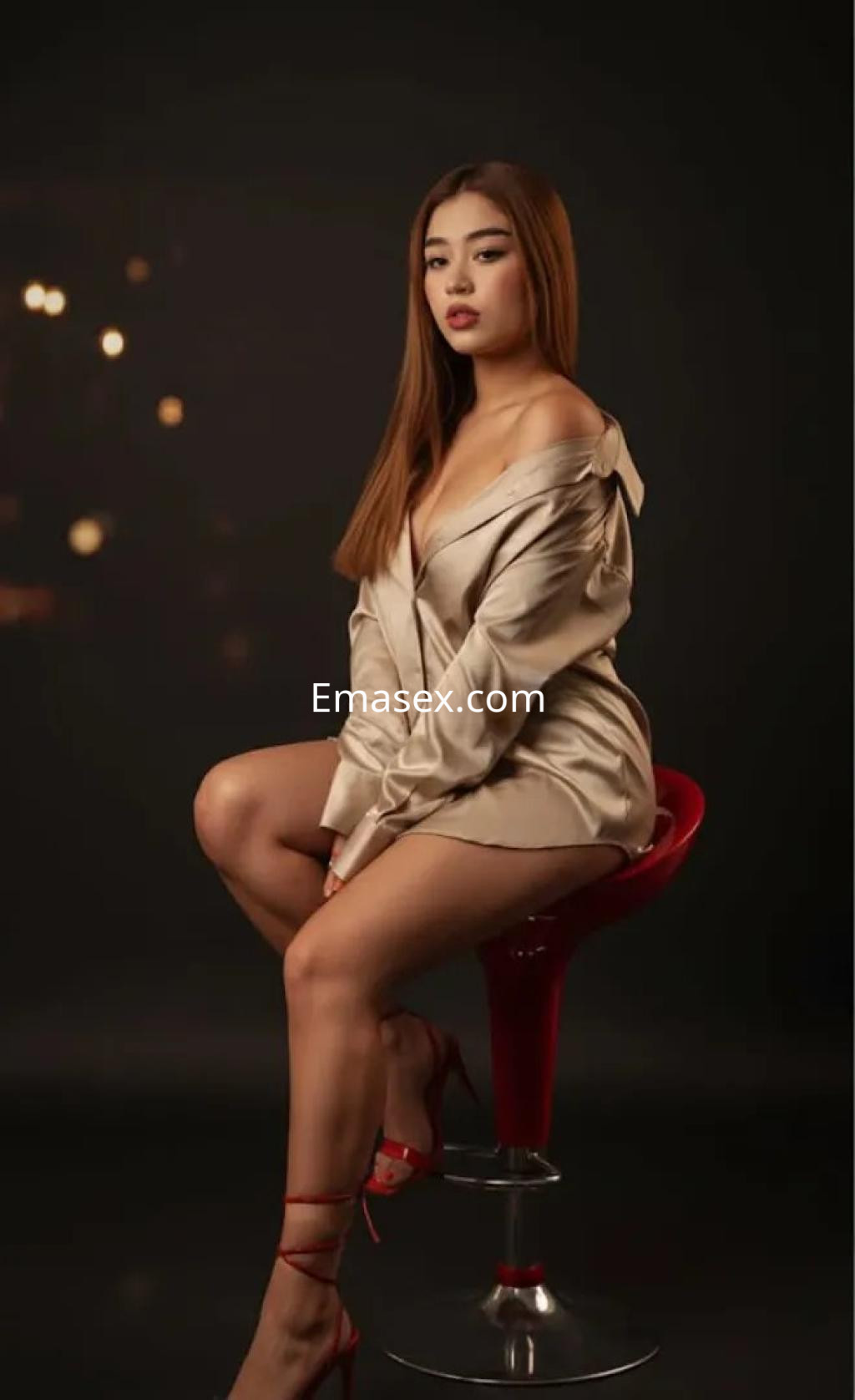 La puta en Córdoba lleva una blusa satinada y posando sentada en una silla roja con fondo oscuro.