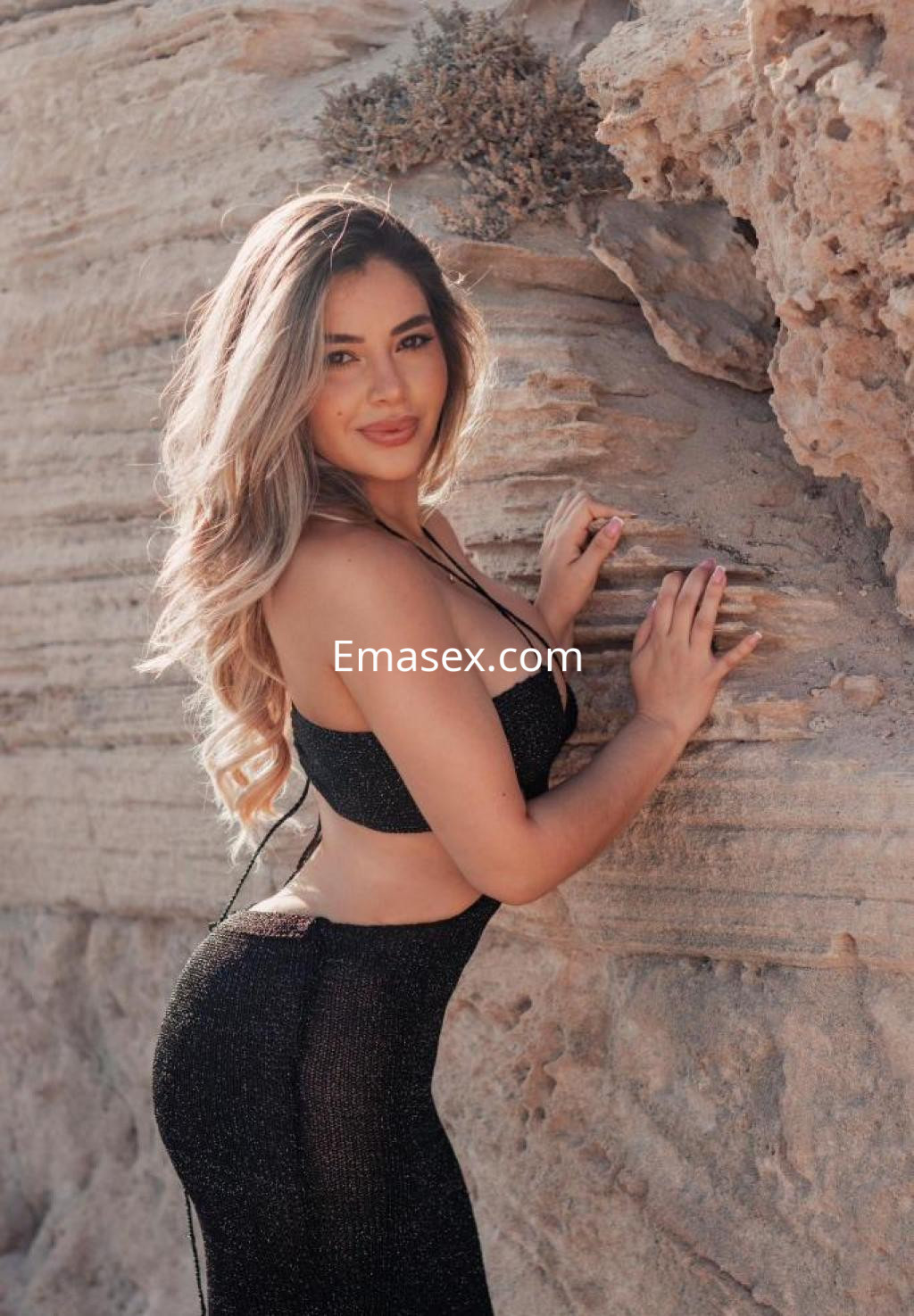 Puta con cabello largo y ondulado, viste un vestido negro ajustado y posa junto a unas rocas en Girona.