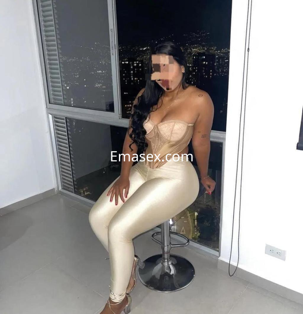 Escort en Girona, sentada en un taburete con un top claro y pantalones ajustados brillantes, mirando hacia la ciudad.