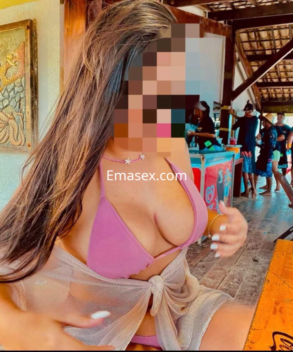Puta con bikini morado y pareo claro, sentada en un bar al aire libre en Oviedo, sonriente y relajada.