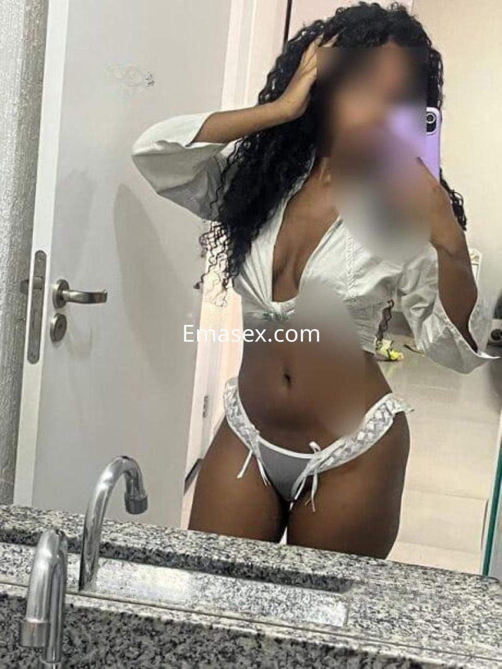 Puta posando en un espejo con cabello rizado y largo, vistiendo una blusa blanca y lencería ajustada en Santander.