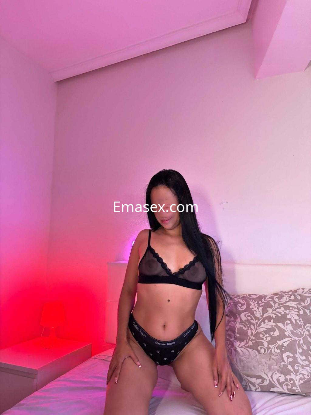 Puta posando sobre la cama en una habitación iluminada con tonos rojos, luciendo lencería negra transparente.