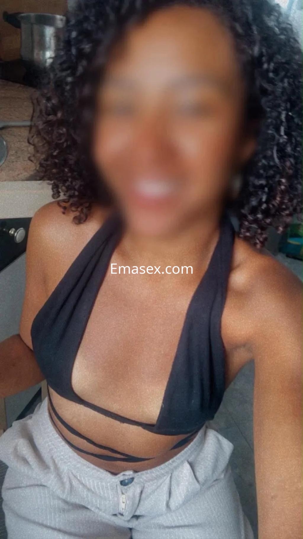 Escort en Bilbao con cabello rizado, viste un top negro y pantalones claros, posando en una cocina.