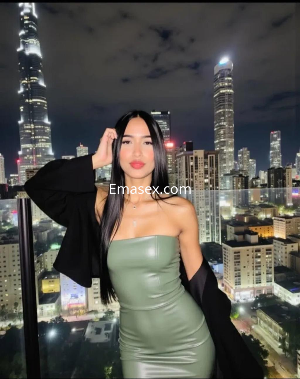 La puta posa en un vestido ajustado verde, con cabello largo y lacio, frente a un paisaje urbano iluminado de Palma de Mallorca.