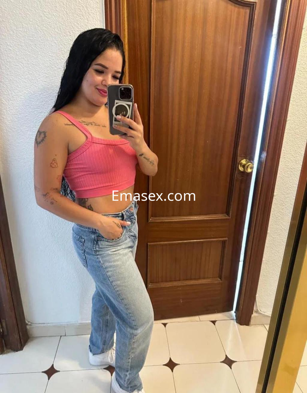 Escort en Córdoba, con cabello largo y oscuro, viste un top rosa y jeans claros, posando frente a un espejo.