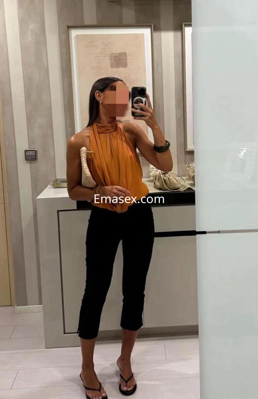 Escort en Lleida con blusa naranja y pantalones negros, posando en un ambiente moderno y luminoso.