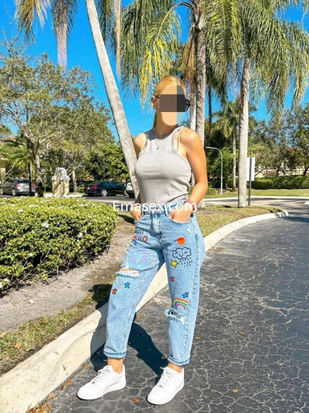 Puta en Lleida viste una camiseta gris y jeans decorados, posando al aire libre entre palmeras.