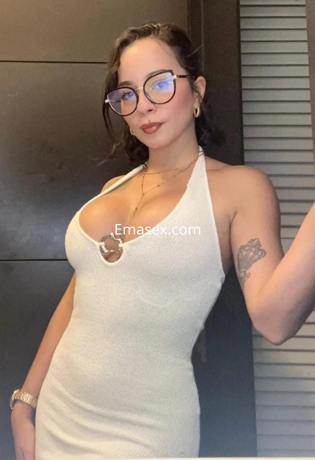 Escort en Málaga luciendo un vestido blanco ajustado y gafas, con cabello corto y un tatuaje visible en el brazo.