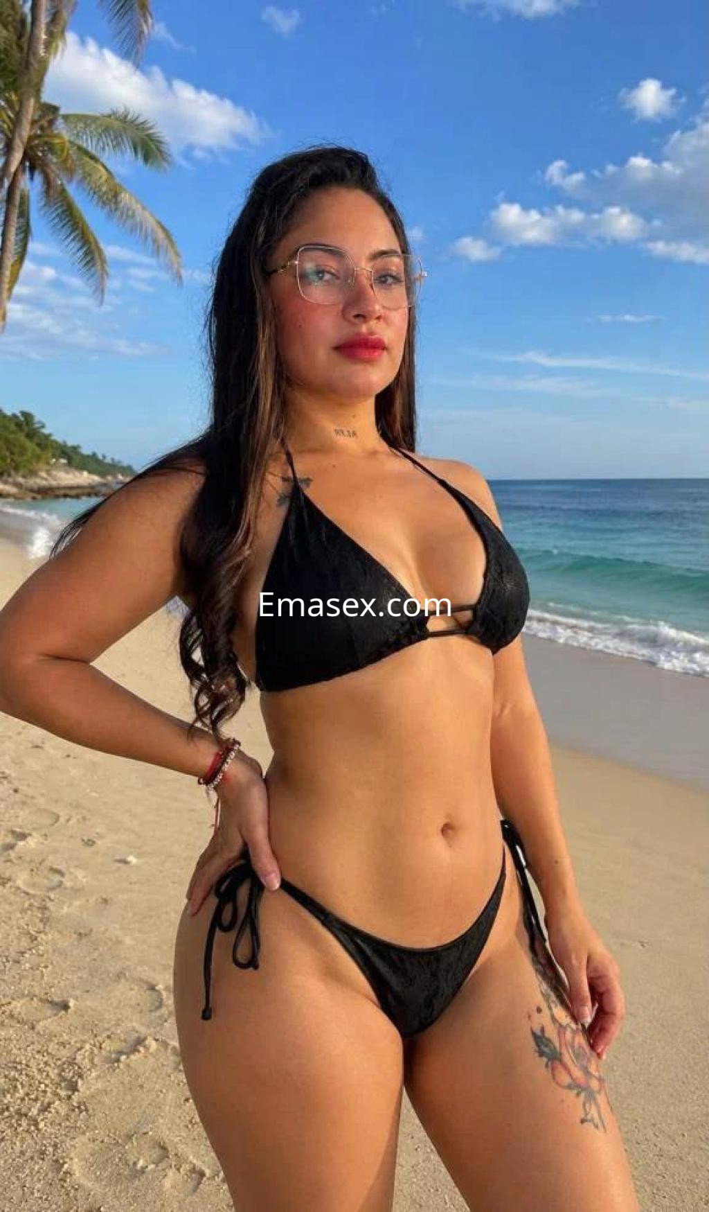 Escort en Málaga con cabello largo, piel bronceada y gafas, posa en la playa con un bikini negro.