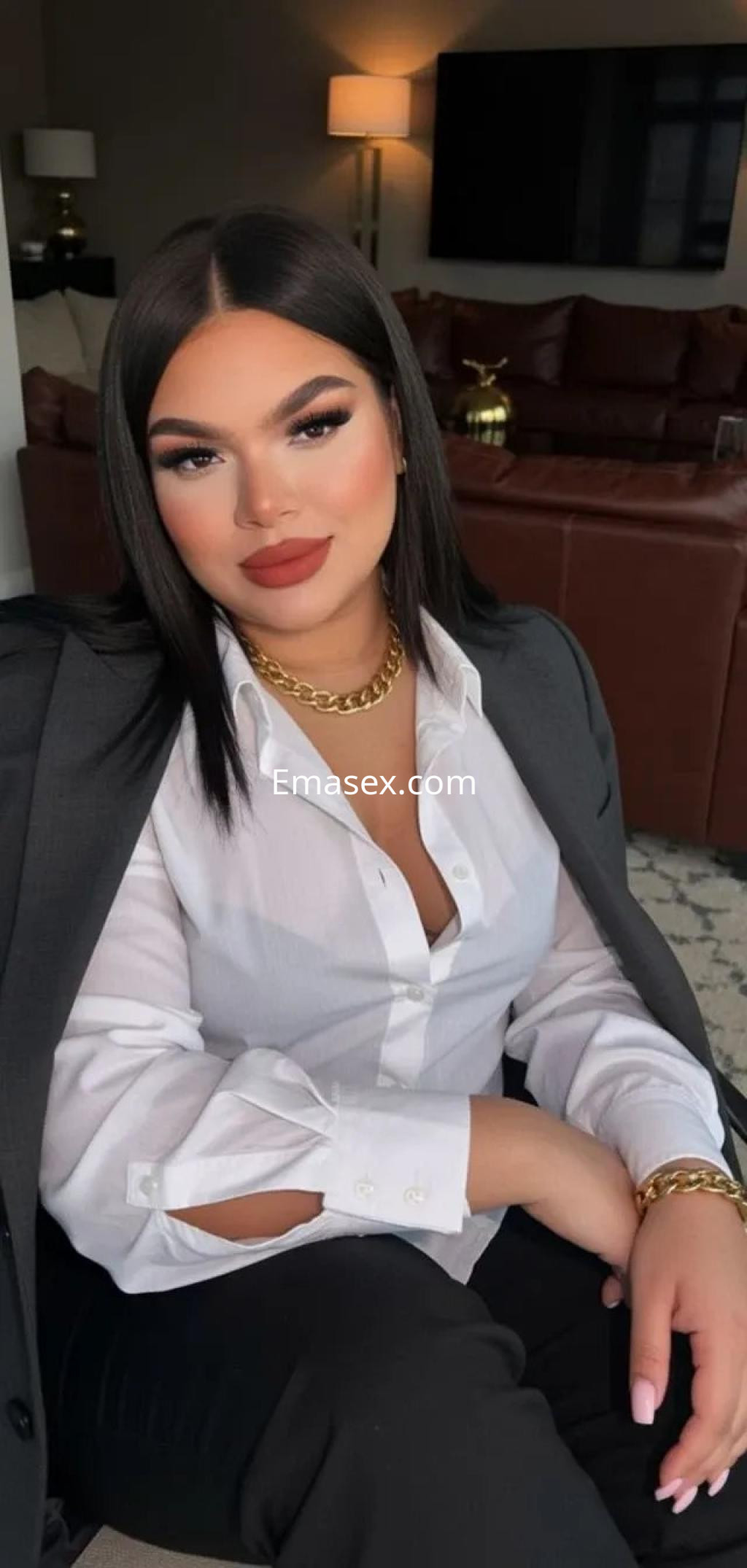 Escort en Murcia con cabello lacio y oscuro, viste una camisa blanca y un conjunto elegante, sonriente en un ambiente acogedor.