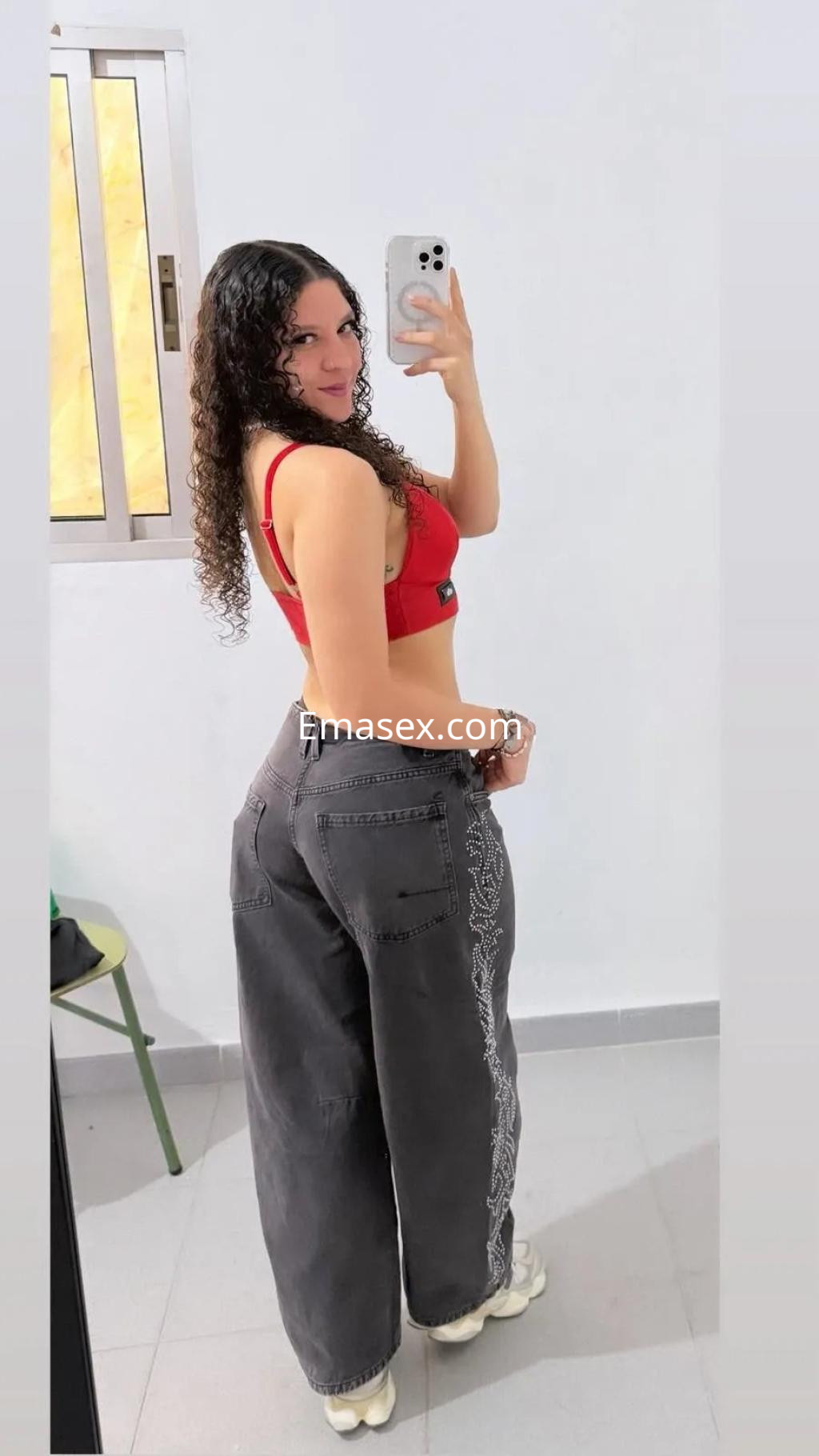 Escort en Valencia mostrando un top rojo y pantalones anchos oscuros, con cabello rizado y posando en un espejo.