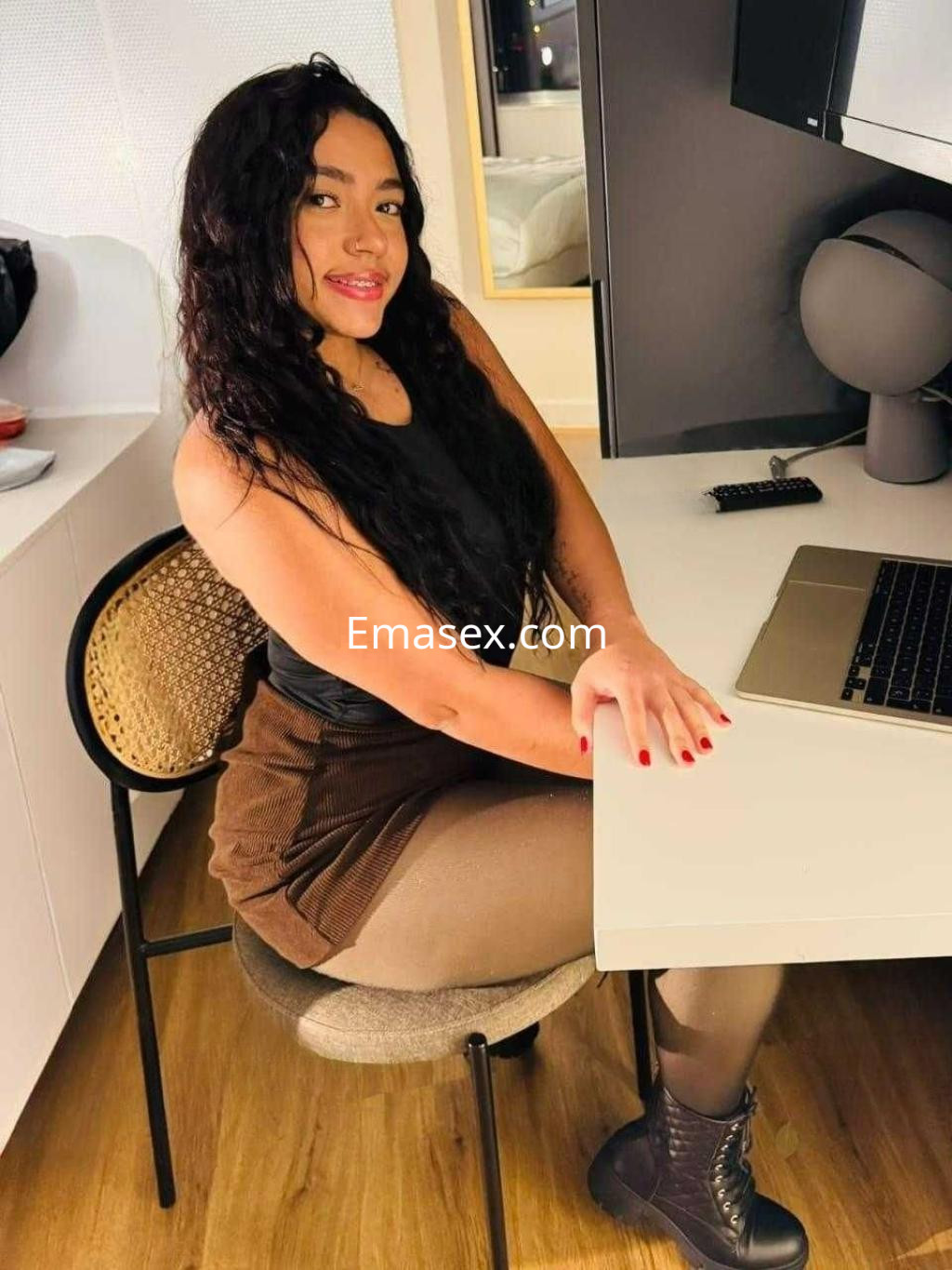 Puta de cabello rizado y oscuro, con blusa negra y falda marrón, posando sentada en una silla frente a una mesa en Valladolid.