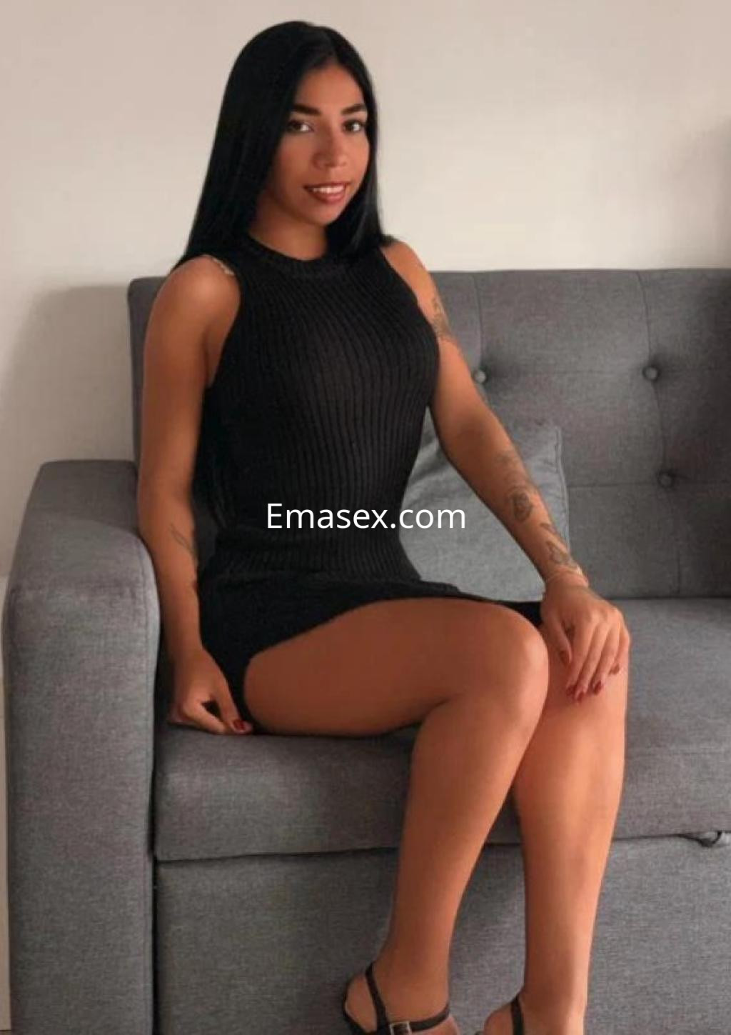 Puta sentada en un sofá gris, luciendo un vestido negro ajustado y sandalias, con cabello largo y lacio en Cádiz.