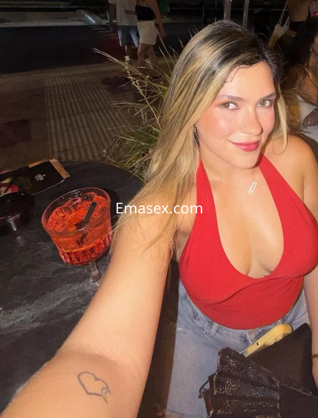 Puta posando con una blusa roja ajustada y jeans, sonriendo en un bar de Palma de Mallorca.