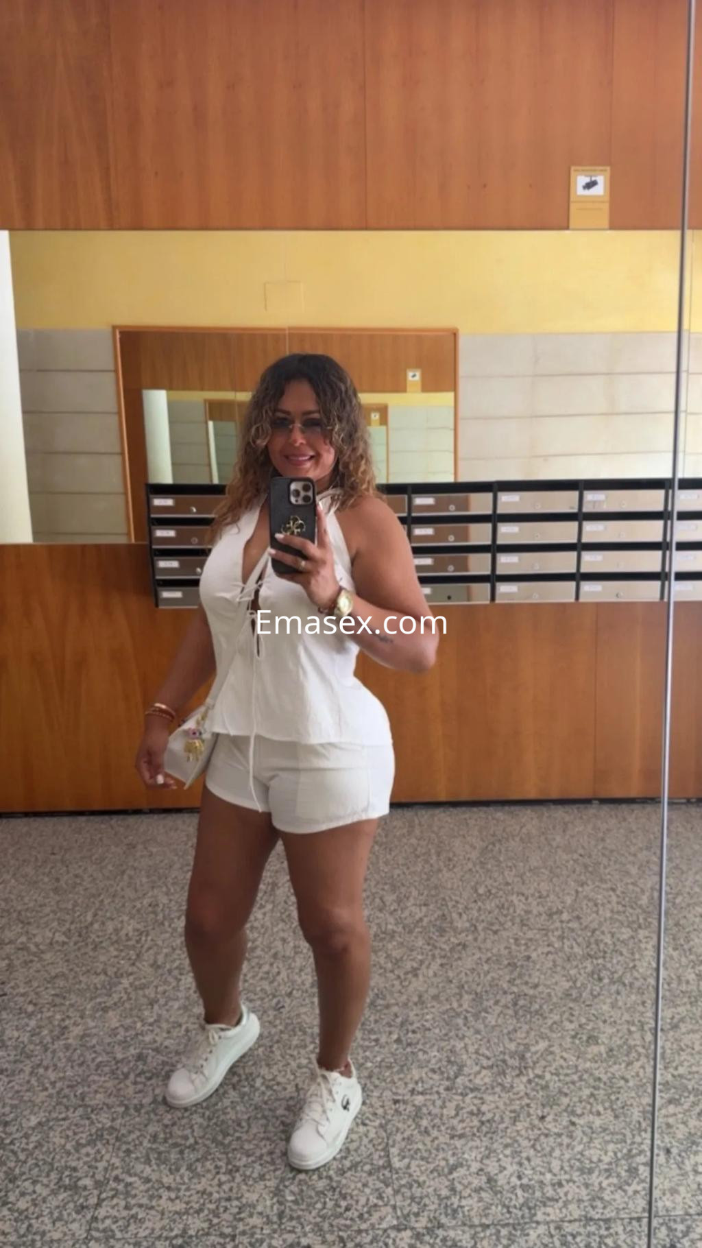 Puta en Murcia con cabello rizado y suelto, luciendo un conjunto blanco y zapatillas deportivas en un espejo.