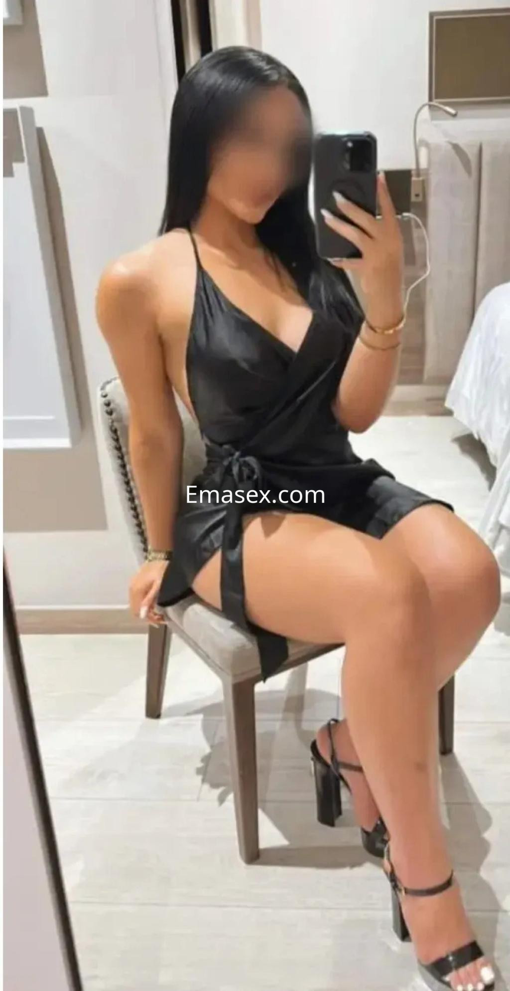 Escort en Murcia sentada en una silla, luciendo un vestido negro ajustado y tacones, con cabello largo y liso.